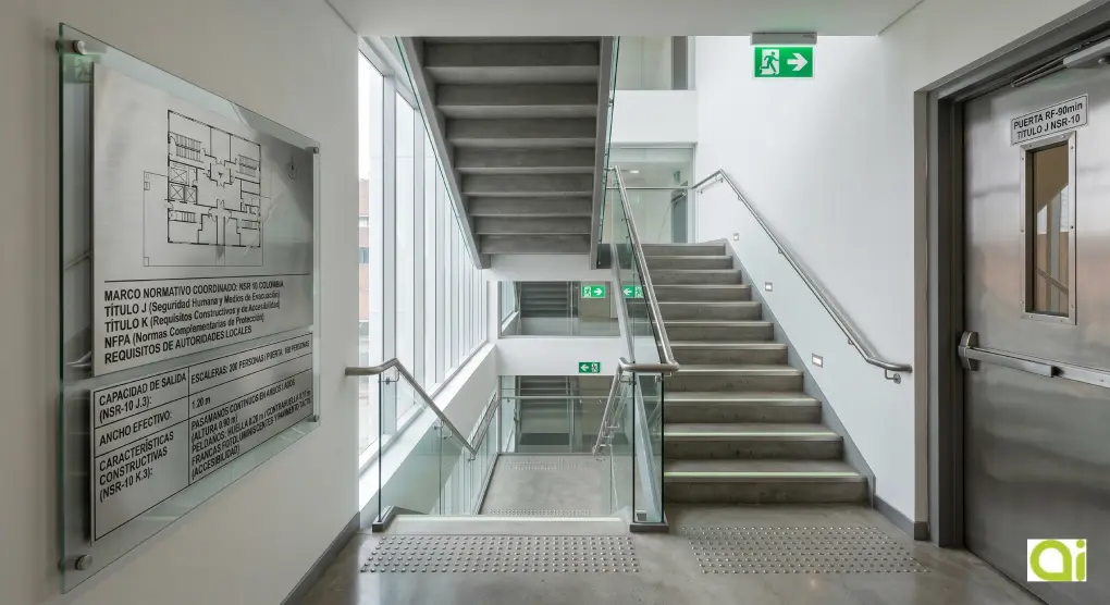 Fotografía técnica que detalla una placa de cumplimiento normativo en una escalera de evacuación en Colombia, conforme a NSR-10 Títulos J y K.